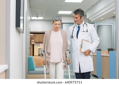 Mature Doctor Helping Old Female Patient In Crutches At The Hospital. Physical Therapist Helping A Woman On Crutches In A Medical Clinic. Professional Doctor And Senior Patient Walking In Hallway.