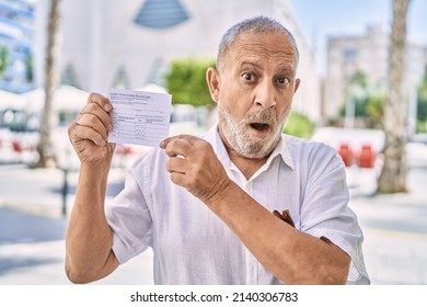 Mature Doctor Man Holding Covid Record Card In Shock Face, Looking Skeptical And Sarcastic, Surprised With Open Mouth 