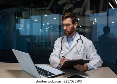 Mature Doctor Working On Paper Work Inside A Shabby Office Building, Man In Medical Coat Using Laptop Filling Out Medical Documents Concentrating And Thinking