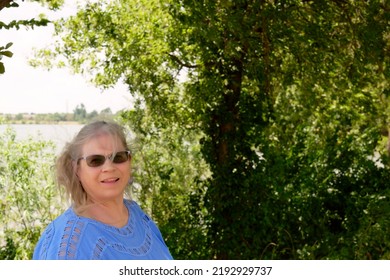 Mature, Elderly Caucasian Woman Standing Next To A Tree With A Lake In The Background.