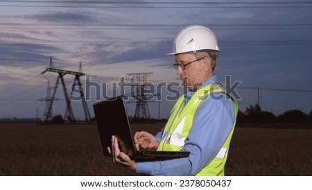 Mature electrician with laptop inspects network of powerlines in evening field. Engineer conducts diagnostics of power generation substation facilities with laptop in sunset country field