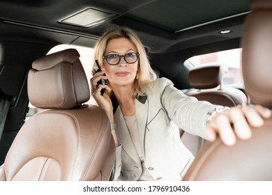 Mature elegant female with blond hair talking on smartphone on backseat of taxi cab and looking through window of driver during traffic jam - Powered by Shutterstock