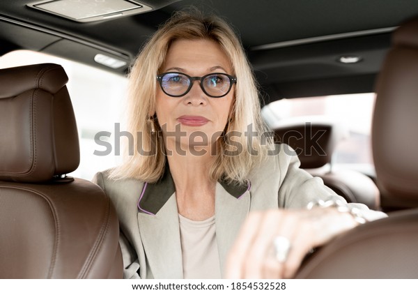 Mature elegant female with blond hair looking at\
you while sitting on backseat of taxi cab between seats and waiting\
for driver