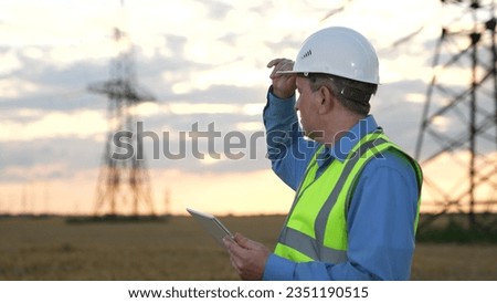 Mature engineer inputs data on tablet and looks back at power transmission line