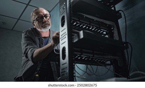 Mature engineer inserts the cable into the communications equipment, uses tablet computer. Male system administrator works in server room. Local area network or web server maintenance. Tech support.