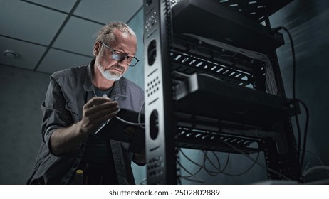 Mature engineer inserts the cable into the communications equipment, uses tablet computer. Male system administrator works in server room. Local area network or web server maintenance. Tech support.