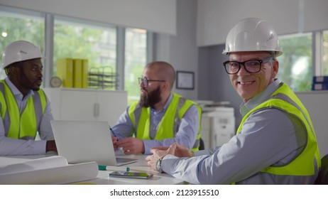 Mature engineer smiling at camera sitting at desk with colleagues. Diverse professional construction workers brainstorming with laptop and blueprints in modern office