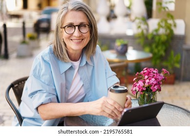 Mature European grey hair charming woman drink coffee using digital tablet sitting at table of street cafe. Mature silver hair woman working outdoors city cafe, wearing white t-shirt and blue shirt - Powered by Shutterstock