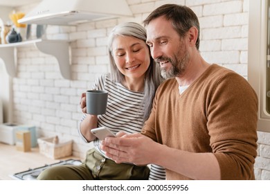 Mature family middle-aged wife and husband couple using smart phone watching videos, social media, reading news on cellphone at home in the kitchen together. - Powered by Shutterstock