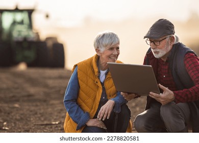 Mature farmers man and woman crouching in field and looking at laptop while tractor plowing in background - Powered by Shutterstock