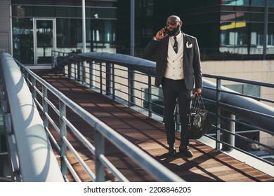 A mature fashionable black man entrepreneur is talking on the phone while passing on an overhead passage; an African bearded businessman is phoning while standing on the pedestrian bridge with his bag - Shutterstock ID 2208846205
