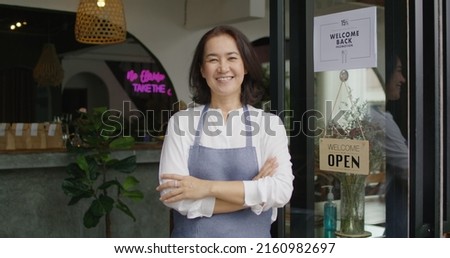 Mature female adult asia people open small bar pub look at camera enjoy laugh work with pride stand at modern SME store. Casual apron chef clerk staff in food or drink cafe coffee shop after pandemic.