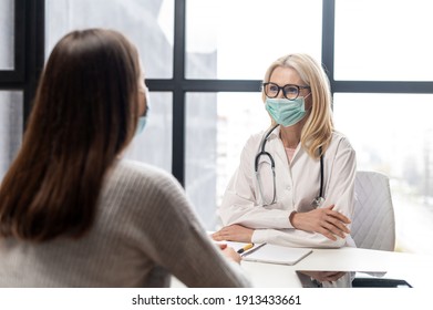 Mature Female Blonde Doctor Wearing Glasses, Uniform, Stethoscope, And Protective Face Mask Giving Advice To A Sick Young Woman Patient At The Hospital, Safety, Social Distancing And Hygiene Concept