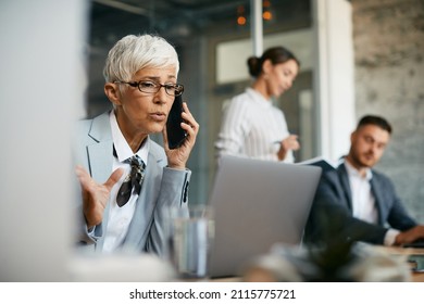 Mature Female CEO Communicating With Someone Over Cell Phone While Working On Laptop In The Office. 