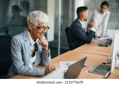 Mature Female CEO Working On Laptop At Corporate Office. Her Colleagues Are In The Background.