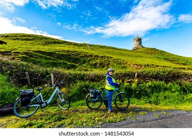 Mature female cyclist taking a break with Doonagore Castle tower in the background, looking at camera, wearing reflective vest and helmet, Wild Atlantic Way, sunny day in County Clare, Ireland - Powered by Shutterstock