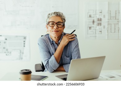 Mature female designer looks at the camera in her office, confident in her expertise and style of creativity. Senior design professional using a laptop and a graphics tablet to put her skills to work.