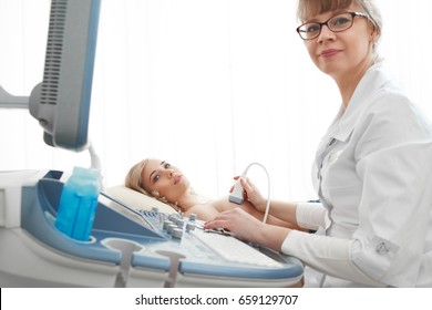 Mature female doctor looking to the camera while examining her female patient performing ultrasound scanning on breast sonography mammologist gynecology.