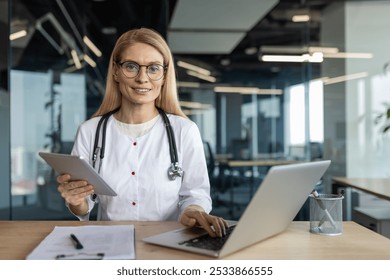 Mature female doctor in office using laptop and tablet for telemedicine. Professional healthcare setting highlights expertise in medical technology. Stethoscope adds clinical touch. - Powered by Shutterstock