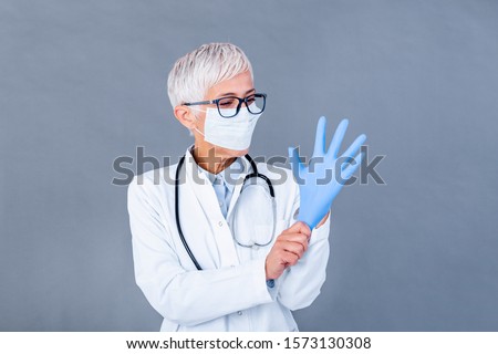 Mature female Doctor putting on protective gloves and medical protective mask, isolated on background. Doctor putting on sterile gloves