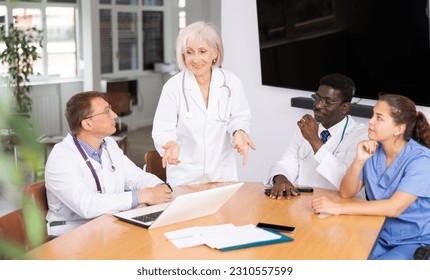 Mature female doctor speaks at meeting of colleagues and tells reports on condition of patients. Medical workers and colleagues participate in discussion and make suggestions and amendments - Shutterstock ID 2310557599