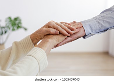 Mature Female In Elderly Care Facility Gets Help From Hospital Personnel Nurse. Close Up Of Aged Wrinkled Hands Of Senior Woman Reaching To A Male Doctor. Copy Space, Background