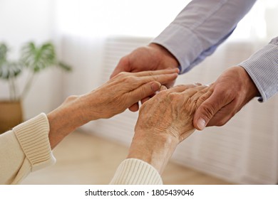 Mature Female In Elderly Care Facility Gets Help From Hospital Personnel Nurse. Close Up Of Aged Wrinkled Hands Of Senior Woman Reaching To A Male Doctor. Copy Space, Background