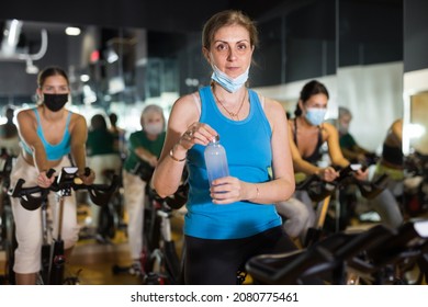 Mature Female In Fase Mask Drinking Water During Cycling Class In Modern Gym