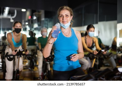 Mature Female In Fase Mask Drinking Water During Cycling Class In Modern Gym