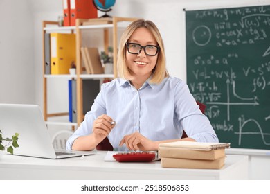 Mature female math teacher sitting at table with laptop and books in classroom - Powered by Shutterstock