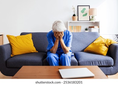 Mature female nurse in blue scrubs covering face with hands feeling overwhelmed sitting on sofa after hard work day in hospital. Sad female healthcare worker resting at home. Health care lifestyle - Powered by Shutterstock