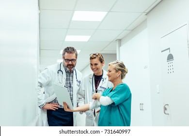 Mature Female Nurse Showing Clipboard To Doctors In Hallway. Team Of Medics Looking At Medical Report While Standing In Hospital Corridor.