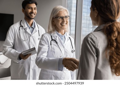 Mature female physician and intern greeting clinic patient, shaking hands, welcoming, start consultation, finishing visit in clinic, give professional medical care and help. Healthcare, communication - Powered by Shutterstock