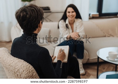 Mature female psychologist in eyeglasses writing down notes during therapy session with smiling young woman