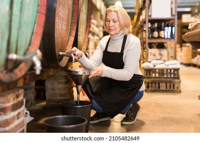 Mature female seller pouring draft wine from wooden barrels to small plastic can in local shop