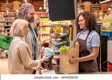 Mature female with smartphone paying for food products in supermarket while standing by her husband in front of happy young cashier - Powered by Shutterstock