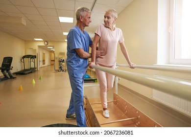 Mature Female Walking Between Parallel Bars In Rehabilitation Room