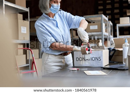 Mature female warehouse worker volunteer wearing face mask packing donations box sealing package in shipping delivery charitable organization. Covid 19 coronavirus donating and volunteering concept.