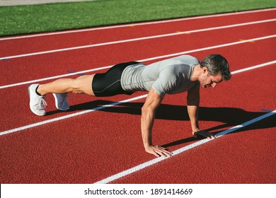 Mature Fitness Man In Black Shorts And Gray T-shirt Doing Push-ups On The Red Running Track. Copy Space. Lifestyle And Sports