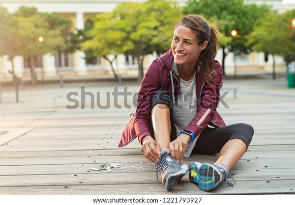 Mature fitness woman tie shoelaces on road.\
Cheerful runner sitting on floor on city streets with mobile and\
earphones wearing sport shoes. Active latin woman tying shoe lace\
before running.