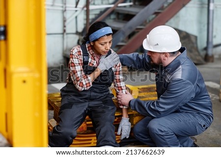 Mature foreman in workwear giving first aid to female subordinate with pain in her shoulder after accident at work in warehouse