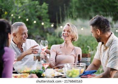 Mature friends having dinner around table in a garden on a summer evening, drinking wine, having fun talking and laughing