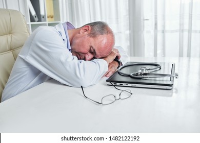 Mature General Practitioner Resting On His Desk With Laptop, Stethoscope And Glasses In Front Of Him