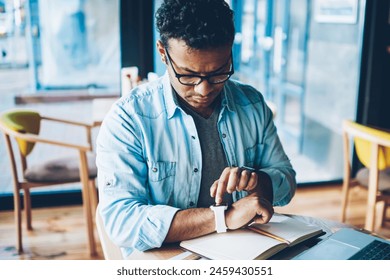 Mature good looking male entrepreneur dressed in casual denim clothes configuring smartwatch while waiting for meeting in coffee shop.Pondering man in eyewear using modern technology for work - Powered by Shutterstock