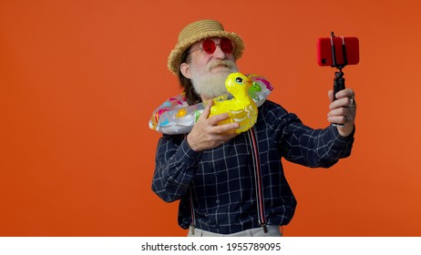 Mature Grandfather Traveler Blogger With Swimming Ring And Inflatable Duck Toy, Taking Selfie On Mobile Phone, Make Video Call Online. Studio Shot. Senior Man On Summer Holiday Vacation, Trip. Tourism