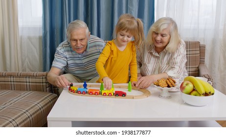 Mature Grandmother, Grandfather With Child Girl Grandkid Riding Toy Train On Railroad On Table In Living Room. Senior Couple Grandparents And Granddaughter Playing Boardgame With Wooden Blocks At Home