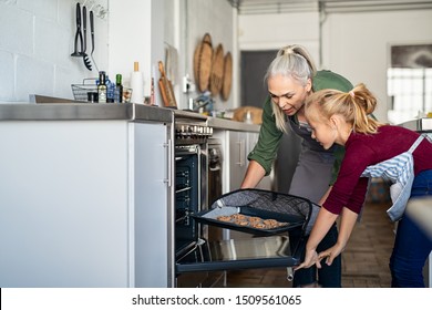 Mature grandmother wearing oven mitts and removing chocolate cookies from oven. Senior grandma taking hot bakeed biscuits out of the oven. Old woman and granddaughter cooking together at home.
