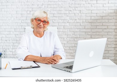 Mature gray haired female doctor is using laptop in hospital office. Senior woman in white formal coat on the white background