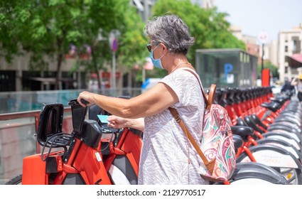 Mature Gray Haired Woman Ready To Rent Electric Bicyle For A City Tour, Holding A Credit Card For Payment. Active Elderly Lady With Backpack Wearing Mask