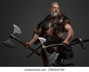 Mature gray-haired bearded Viking warrior in fur and light armor, wielding two enormous axes against a neutral background